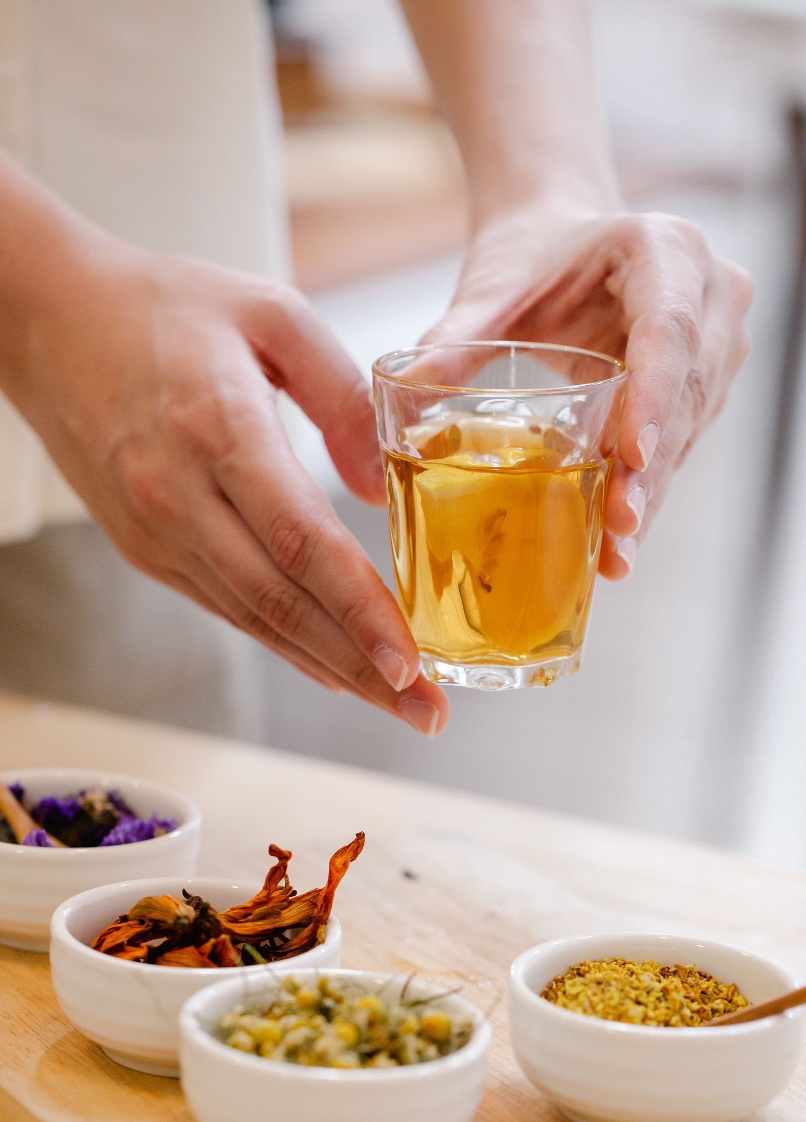 Hand holding a glass of brewed herbal tea beside bowls of dried botanicals